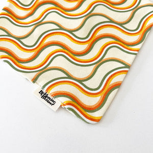 Bohemian Babies made in the USA organic cotton bandana bib. Snap closure. Catch The Wave print. Retro orange, green and mustard waves on an off-white background. Close-upview.